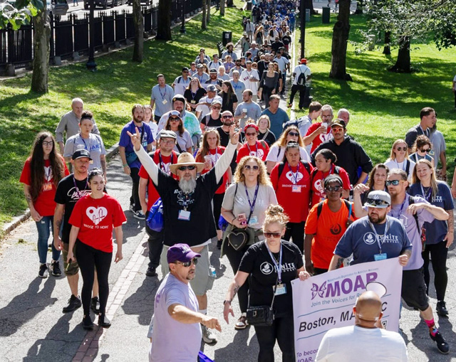 large group of participants in matching t shirts hold MOAR banners and walk through a grassy park